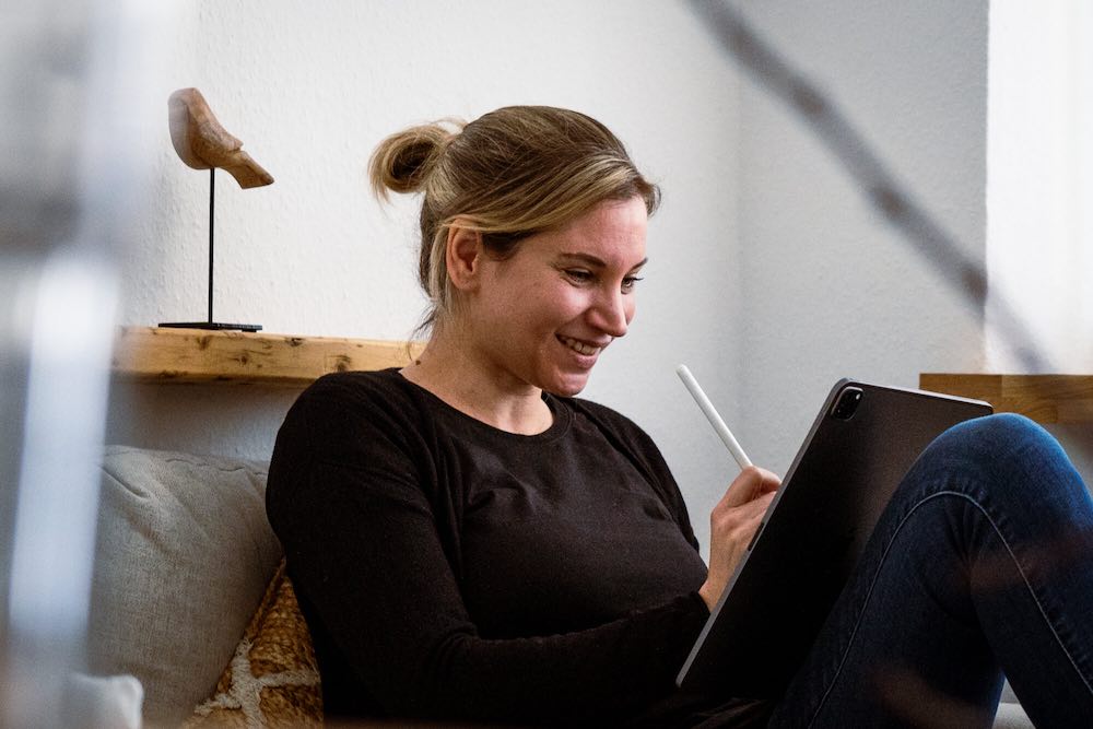 Eva Jentgens sitzt an einer Couch und schreibt lächelnd auf ihrem Tablet.