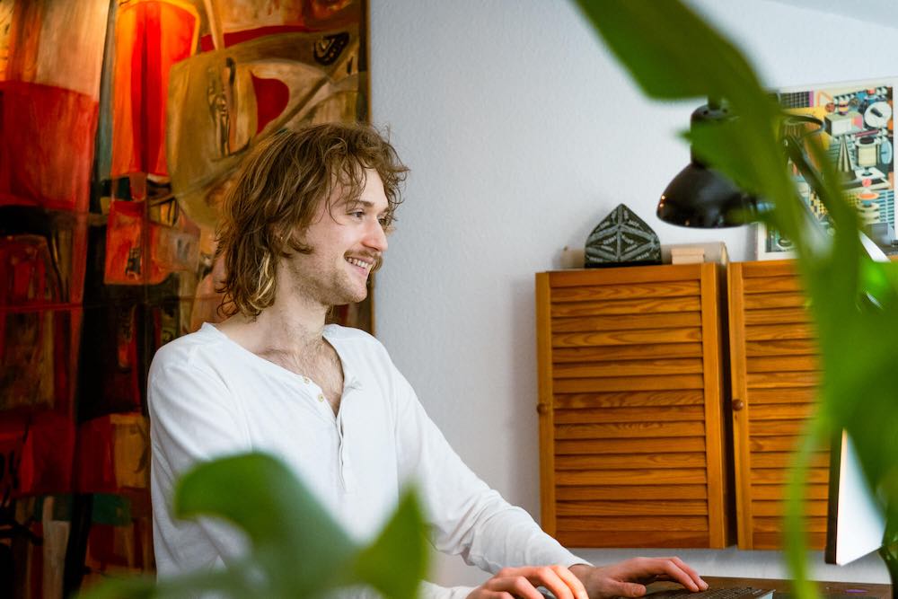 Jano Paetzold steht vor einem großen roten Ölgemälde an einem Stehschreibtisch und schaut lächelnd auf einen Monitor.