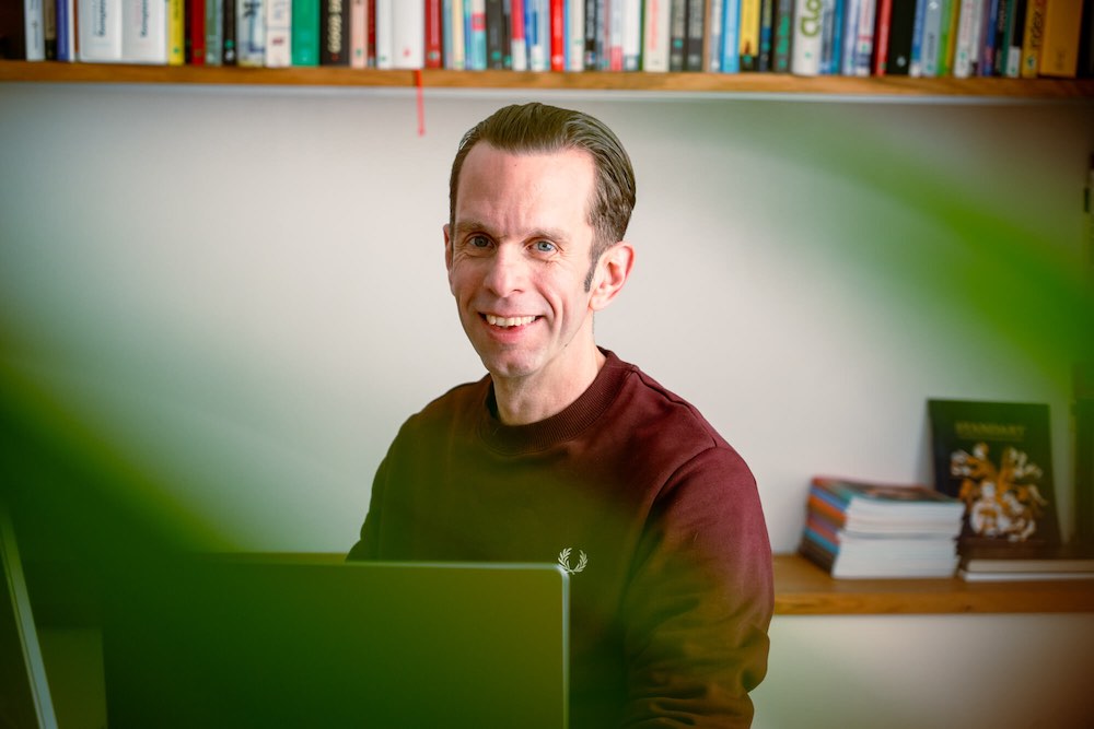 Martin Güthler sitzt vor einem Wandregal mit vielen bunten Büchern und lacht in die Kamera.