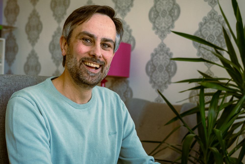 Matthias Pigulla sitzt an seinem Schreibtisch, im Hintergrund eine Pflanze und eine pinke Stehleuchte, unter der er in vielen Besprechungen sitzt.