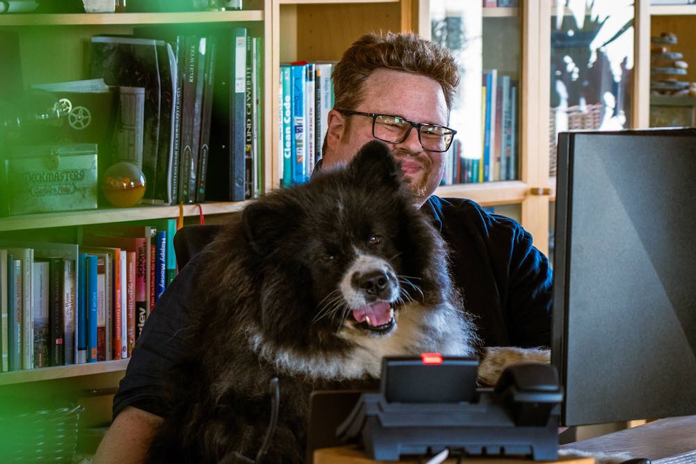 Malte Wunsch und sein Hund Gamma sitzen vor einem Regal mit vielen Büchern und schauen freundlich in die Kamera.