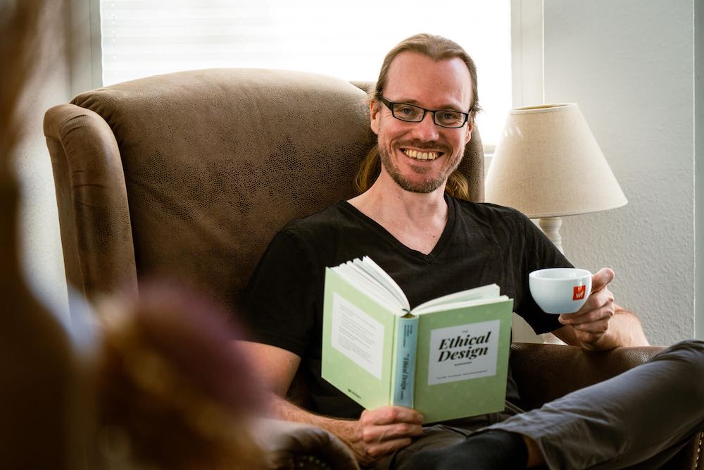 Søren Birkemeyer sitzt in einem brauen Sessel und hält dabei ein Buch und eine Cappuccinotasse in den Händen, dabei schaut er lächelnd in die Kamera