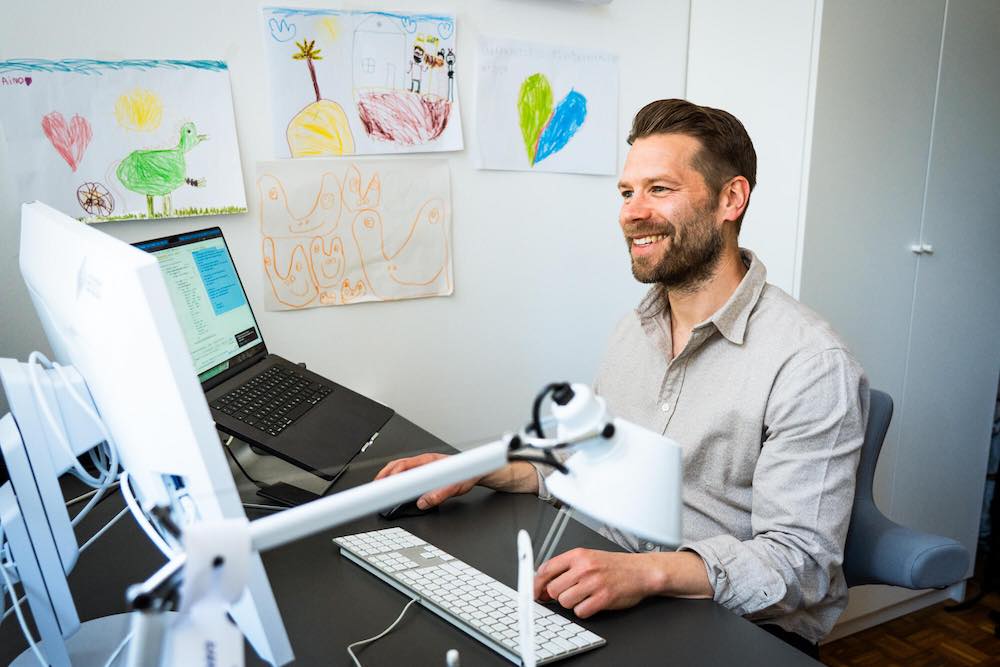 Stefan Hilger sitzt an einem Schreibtisch und schaut lächelnd auf einen Monitor, an der Wand hängen bunt gezeichnete Kinderbilder.